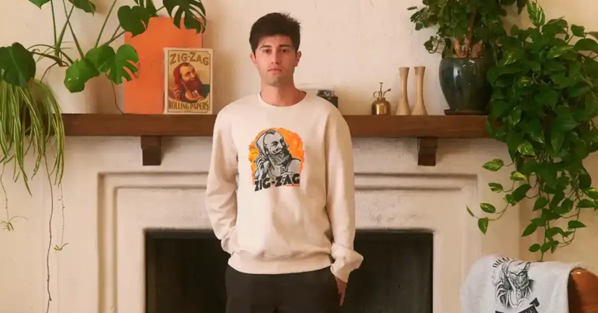 Man wearing a beige Zig-Zag sweatshirt stands in front of a decorated fireplace