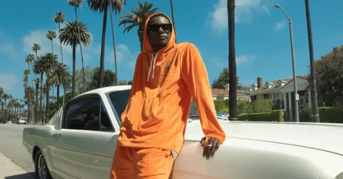 Man in an orange Zig-Zag hoodie and shorts leaning against a white classic car on a palm-lined street