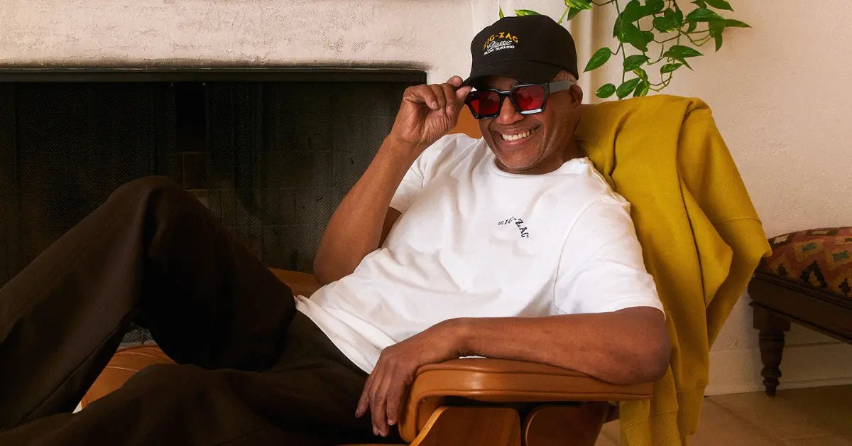 Streetwear clothing style featuring a man lounging in a casual white tee, dark pants, and bold sunglasses indoors