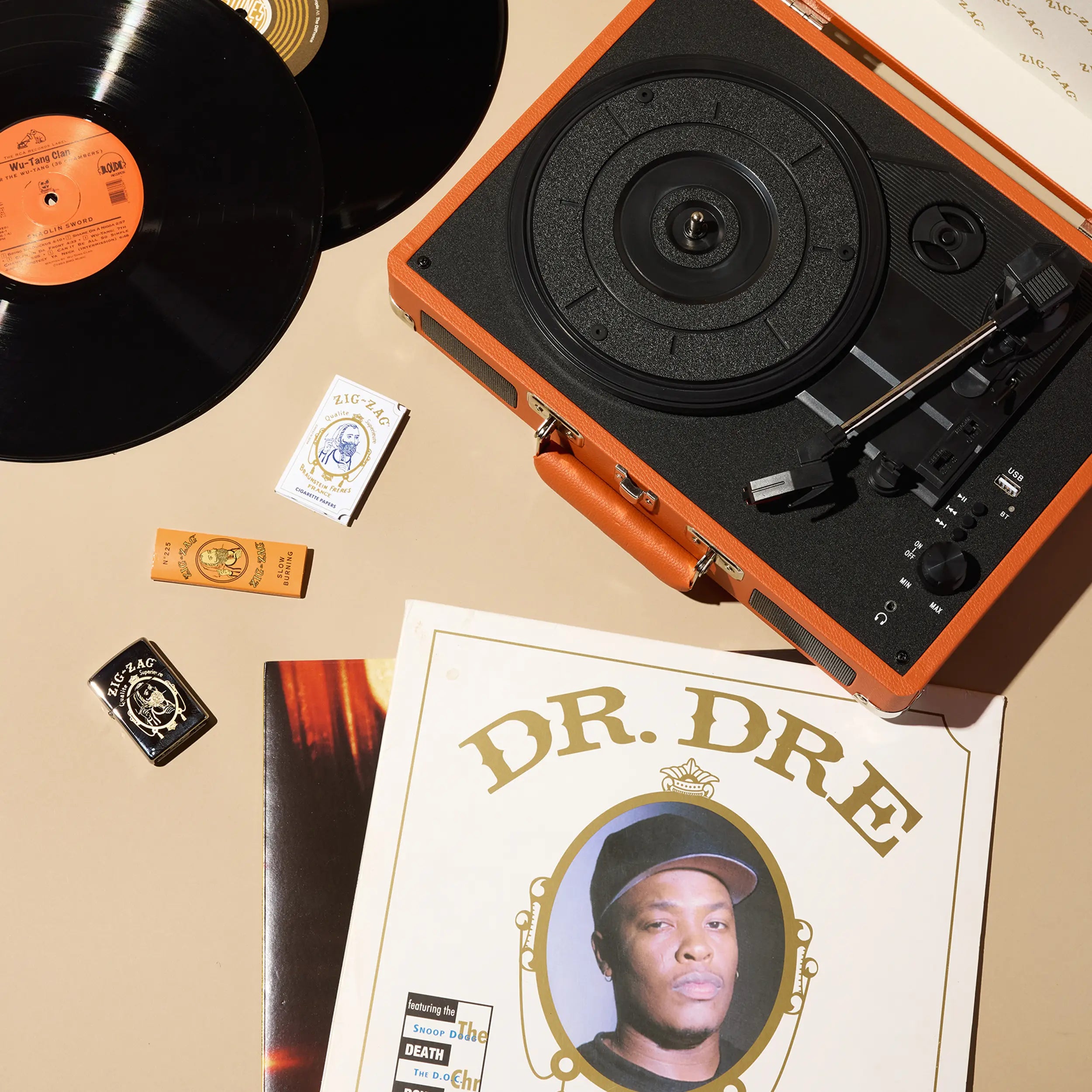 Orange Zig Zag record player surrounded by vinyl records, Zig Zag rolling papers, lighters, and a Dr. Dre album cover.