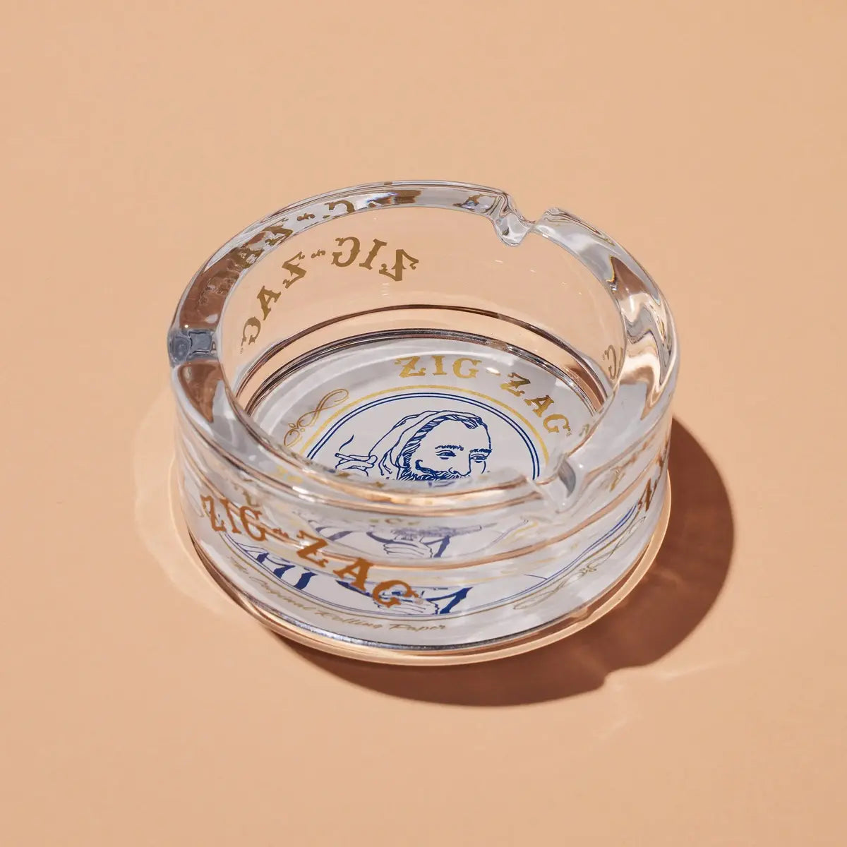 Zig-Zag clear glass ashtray featuring a vintage-style logo design in blue and gold, displayed on a beige surface.