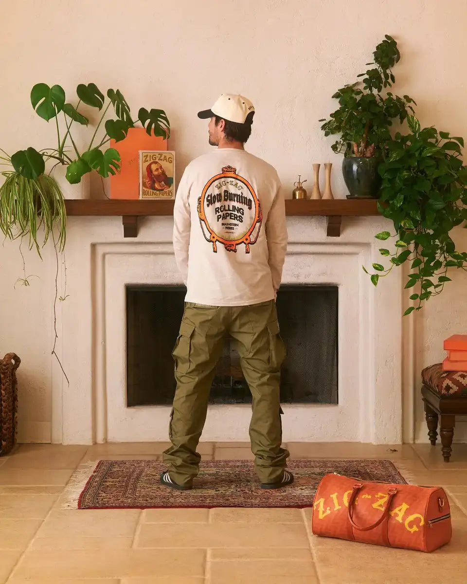 Zig-Zag long-sleeve white shirt with vintage "Slow Burning Rolling Papers" graphic, worn by a person in a cozy room with plants and a branded duffel bag.