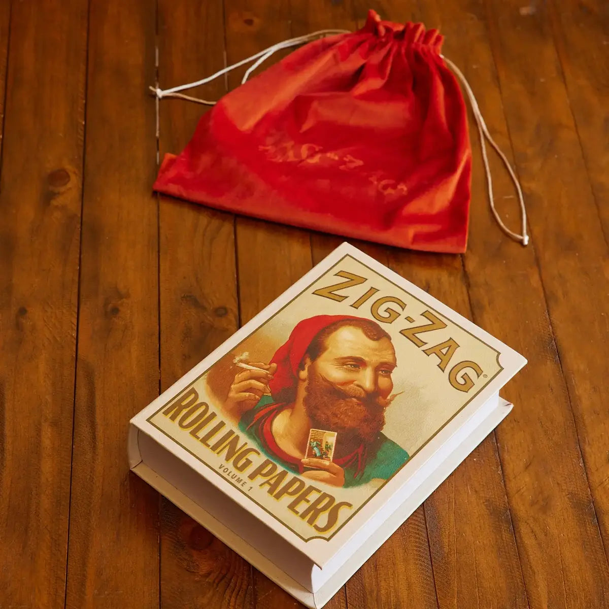 Zig-Zag stash book designed like vintage rolling papers packaging, placed on a wooden surface with a red drawstring bag.