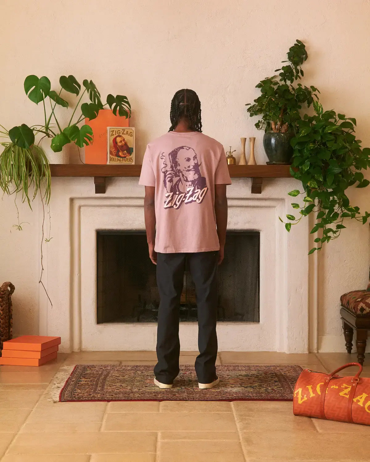 Man wearing a mauve Zig-Zag t-shirt with a large back graphic, standing in a stylish room with plants and decor.