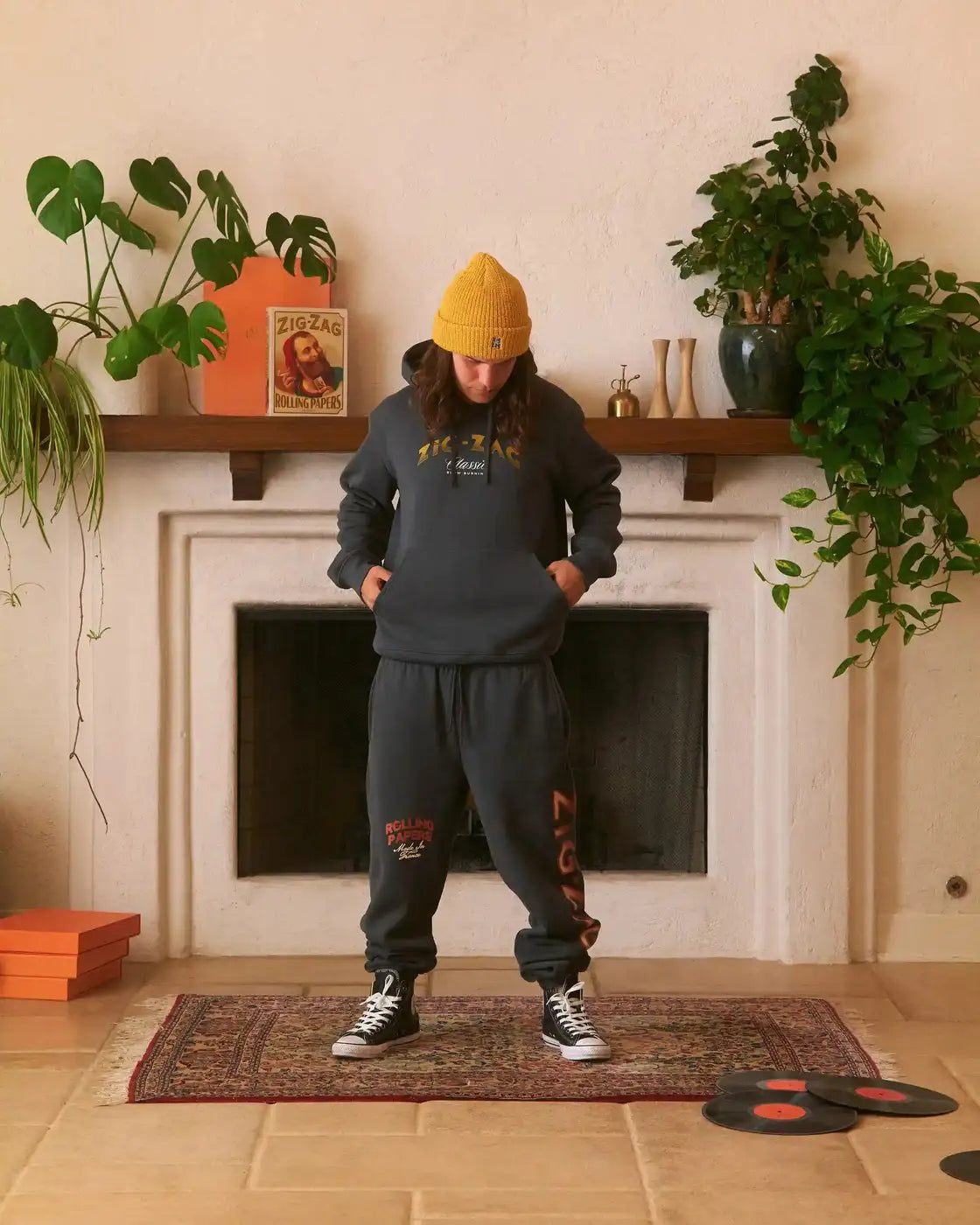 Zig-Zag gray hoodie and sweatpants set with logo graphics, worn by a person in a cozy room with plants and records.