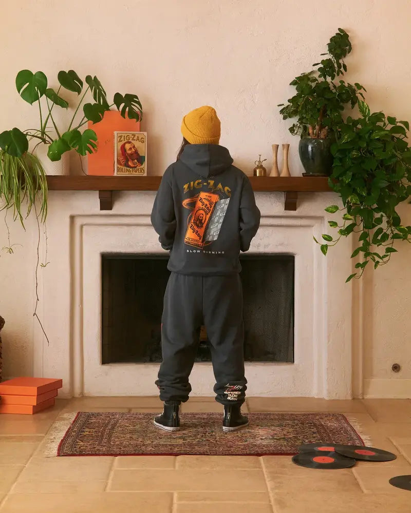Man wearing a black Zig-Zag hoodie with a graphic design, yellow beanie, and pants, standing by a fireplace.