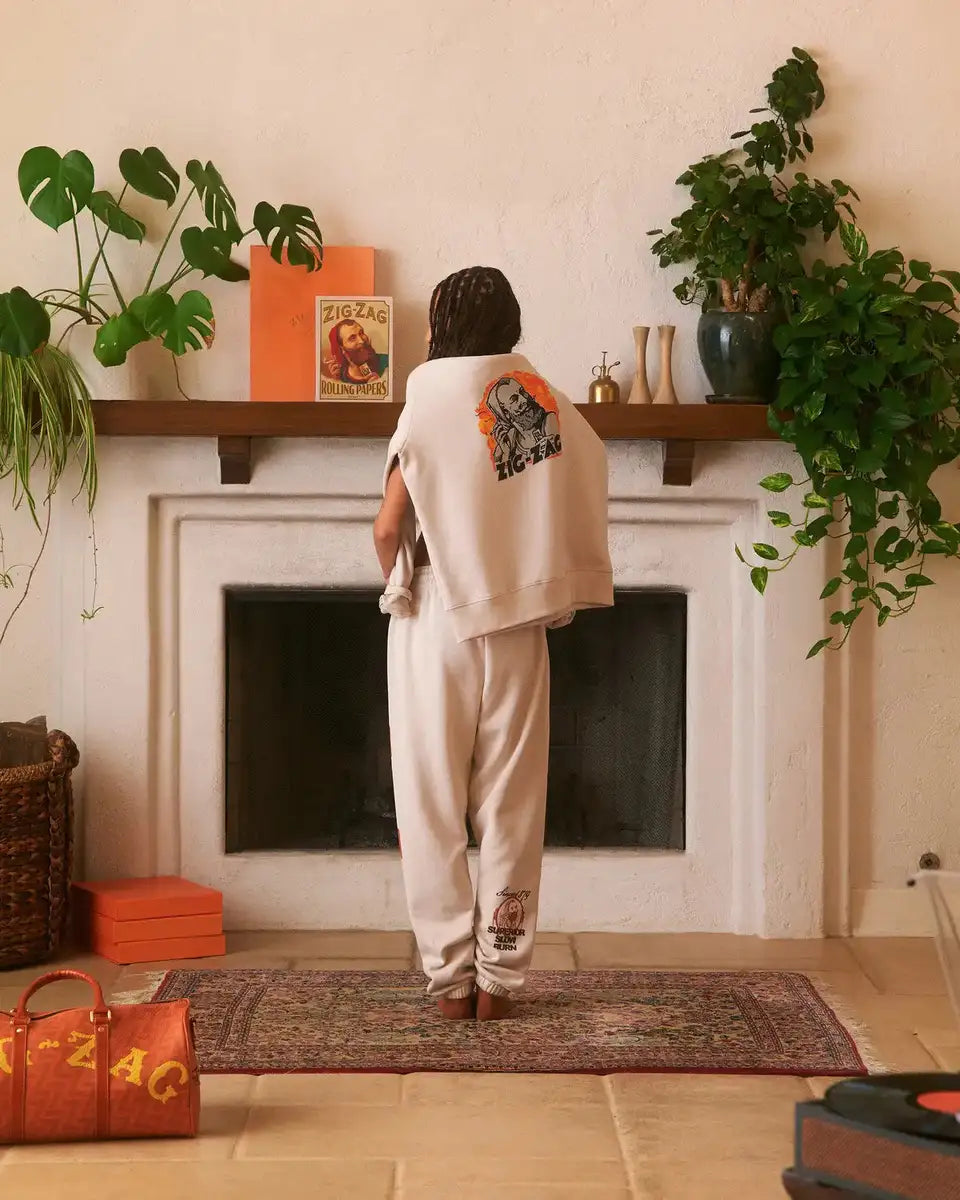Zig-Zag beige sweatpants and matching sweatshirt draped over shoulders, worn by a person in a cozy room with branded decor.