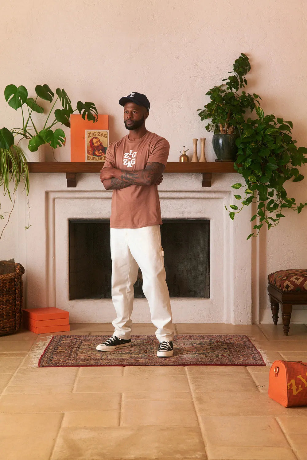 Man wearing a brown Zig-Zag t-shirt and white pants, standing in a cozy room with plants and decor.