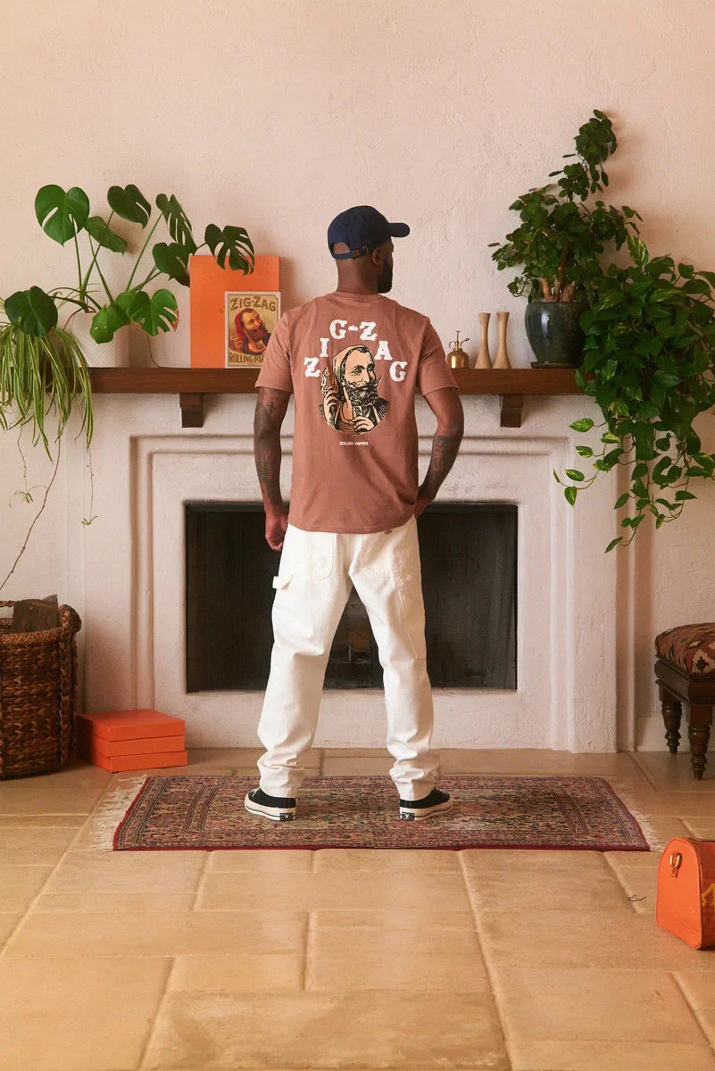 Man wearing a brown Zig-Zag t-shirt with a large back graphic, standing in a stylish room with plants and decor.