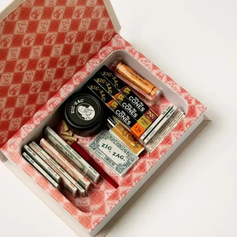 Open Zig Zag stash box filled with assorted rolling papers, cones, a grinder, and accessories arranged in a decorative red interior.
