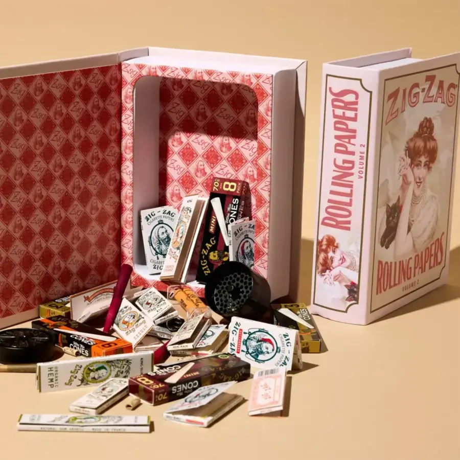 Zig Zag stash box with vintage book design spilled open, revealing a variety of rolling papers, cones, and smoking accessories.