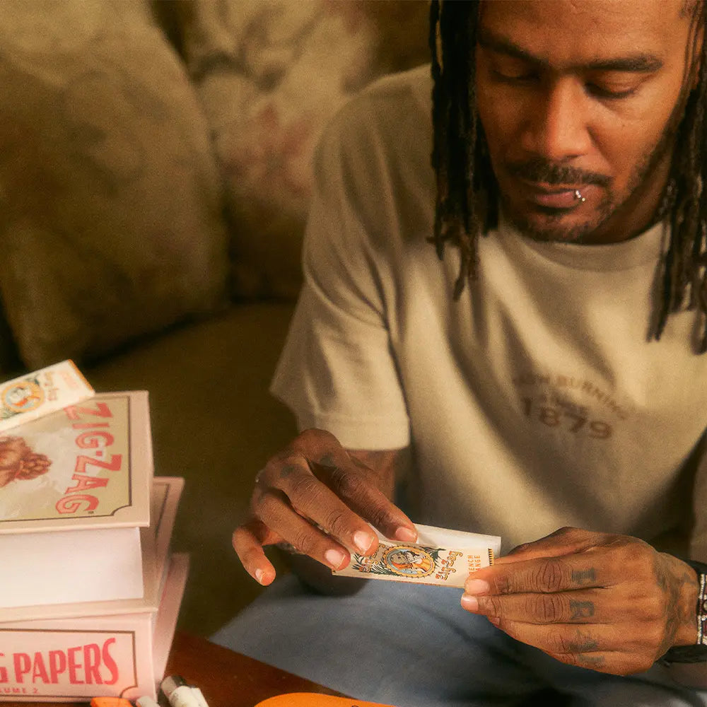 Zig Zag shirt worn by man rolling paper, featuring “Slow Burning Since 1879” text and vintage-style design.