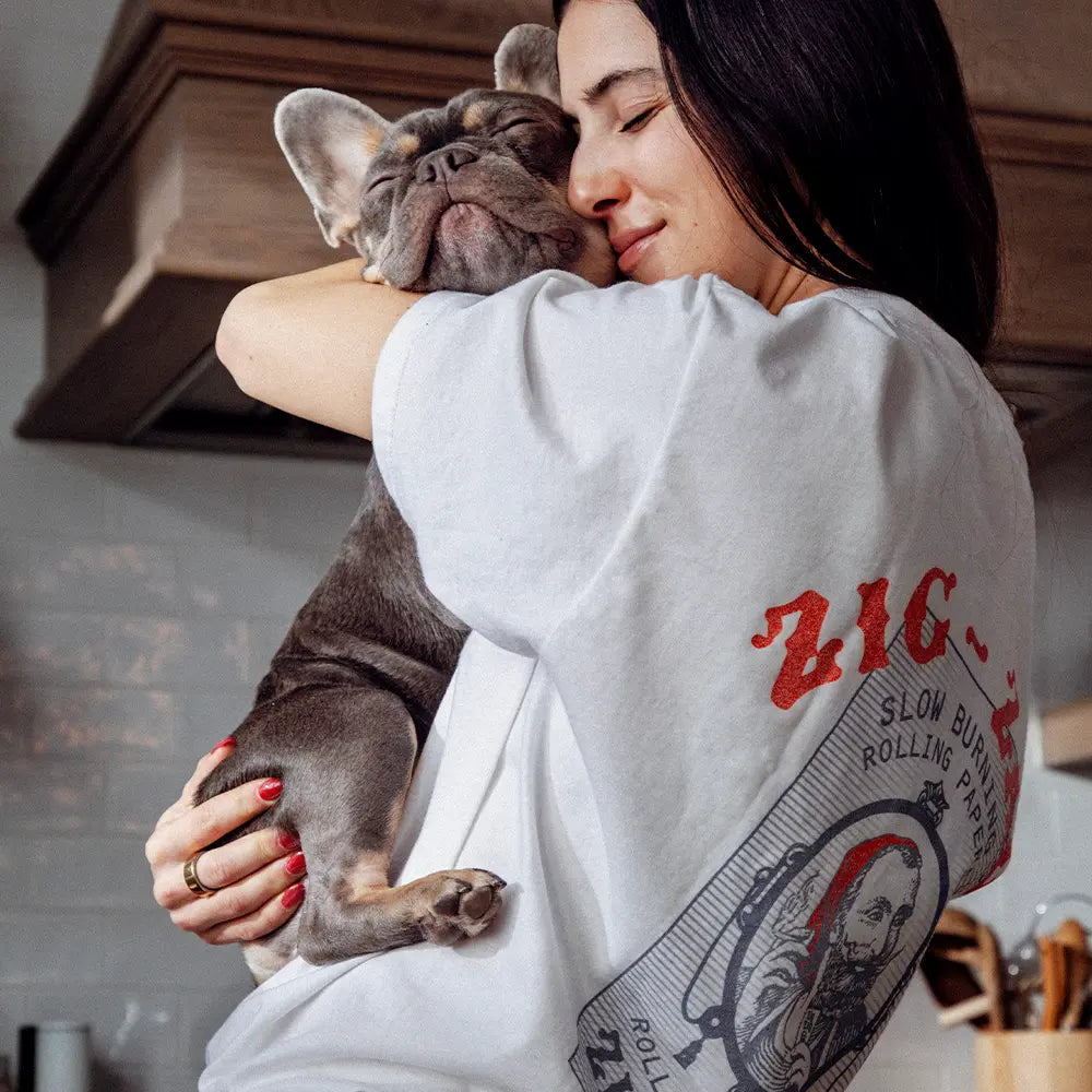 White Zig Zag t-shirt featuring vintage rolling paper artwork worn by a woman cuddling a French Bulldog in a cozy kitchen.