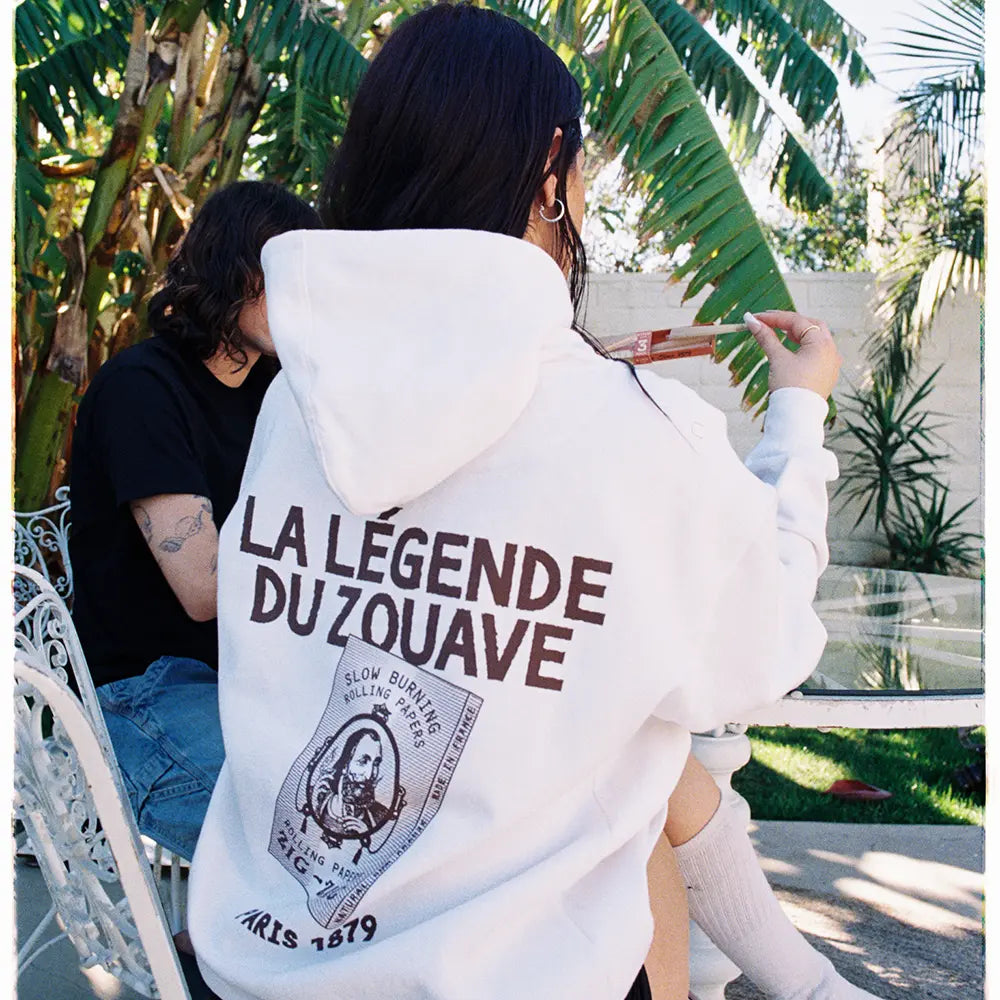 White Zig Zag hoodie worn outdoors featuring bold brown vintage print with “La Légende du Zouave” and rolling paper artwork.