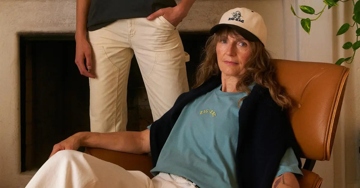 Older woman wearing a Zig-Zag hat and t-shirt seated in a chair, with another person standing behind
