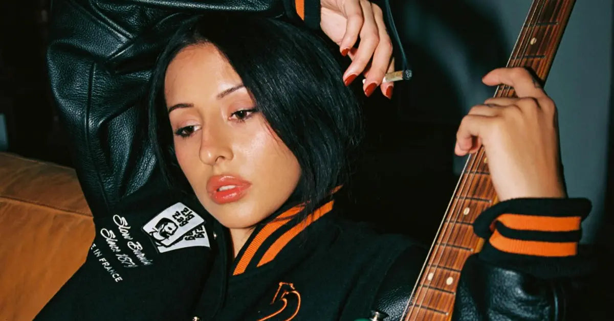 Woman in a black Zig-Zag varsity jacket holding a guitar, lounging with a relaxed expression