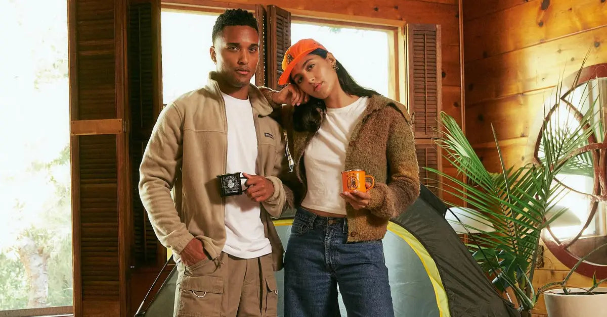 A man and woman in casual attire holding Zig-Zag mugs, standing near a tent in a cozy wooden cabin