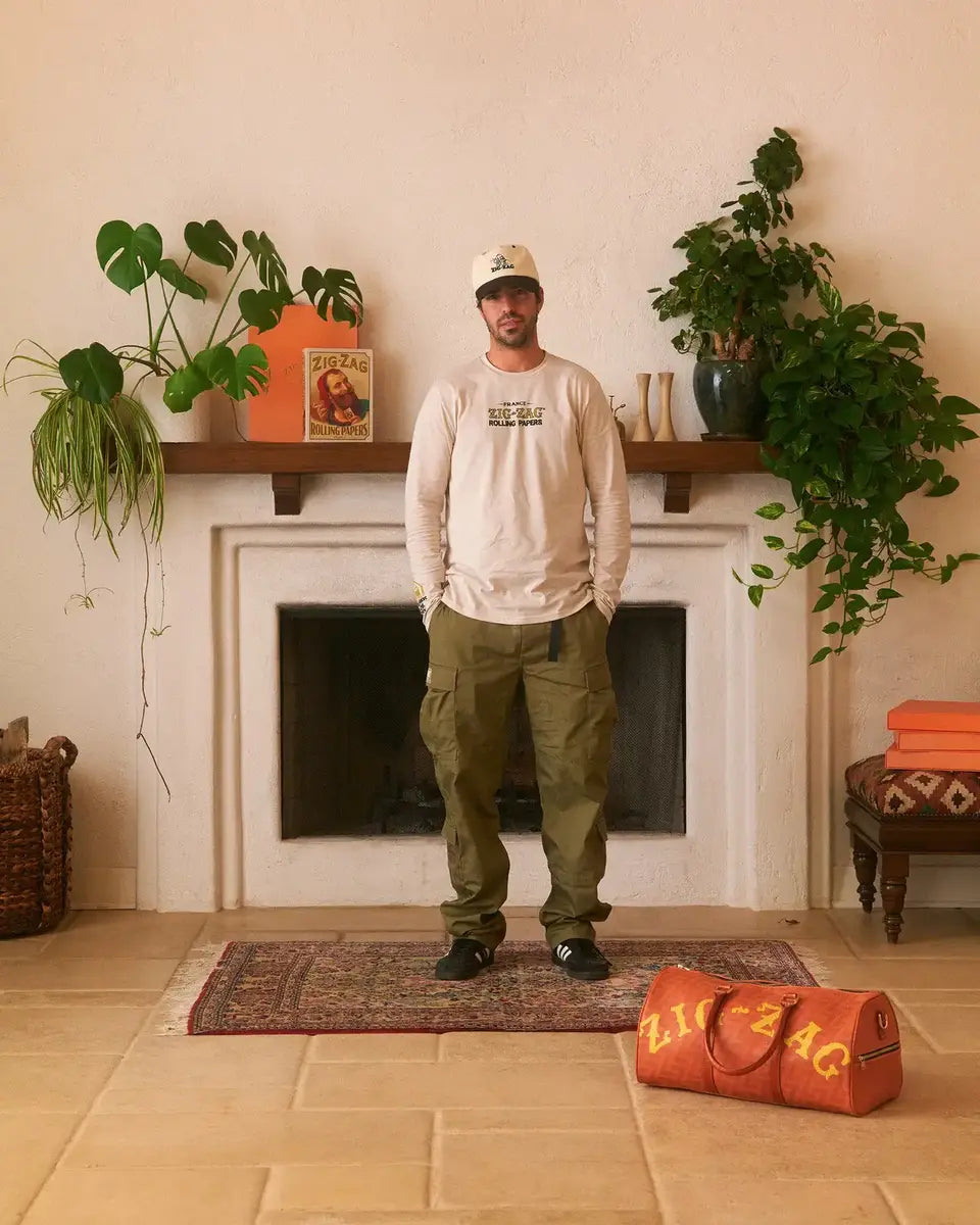 Man in a cream long-sleeve shirt and cap stands in front of a fireplace, next to a Zig-Zag duffel bag.