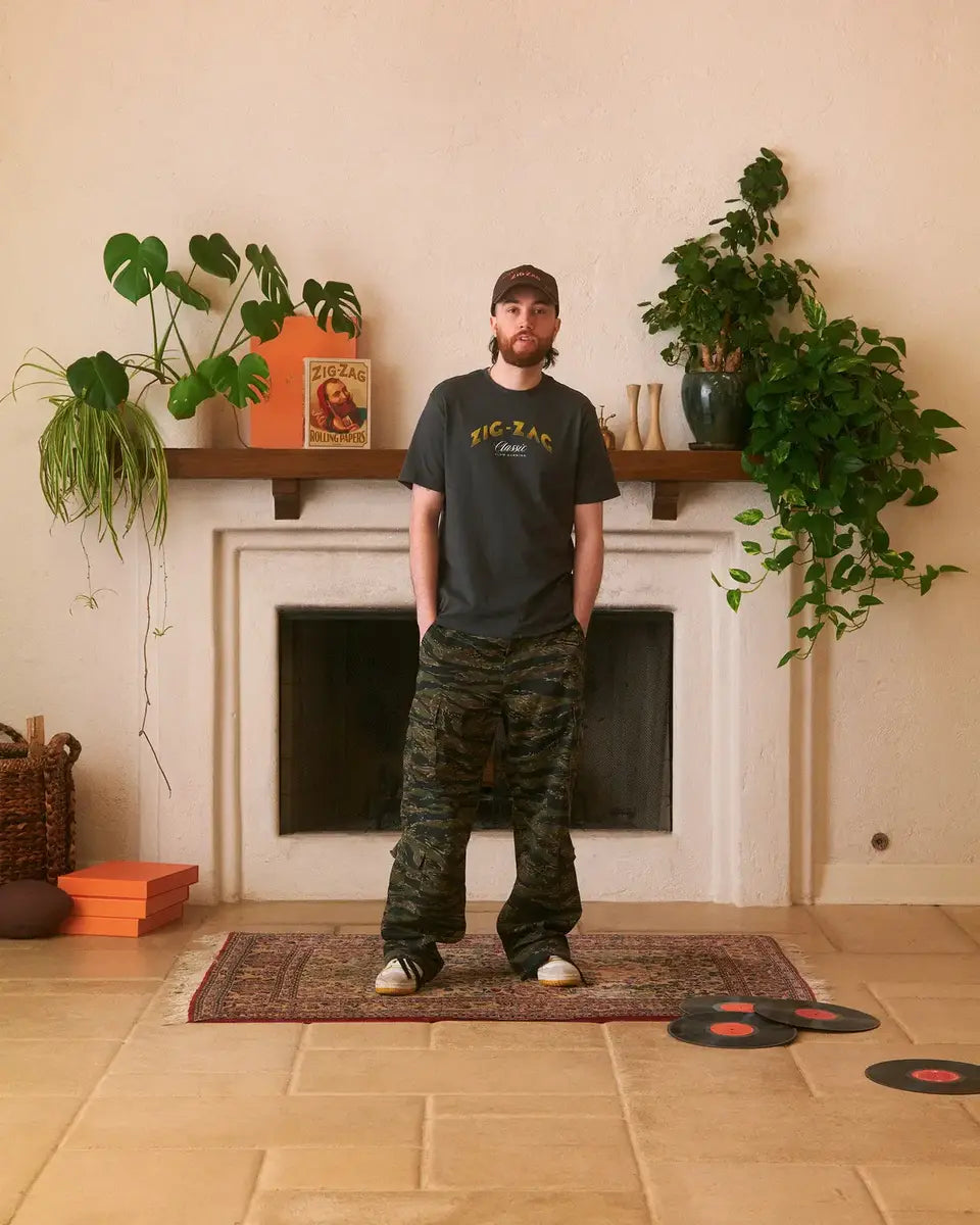 Man wearing a black Zig-Zag T-shirt and camouflage pants stands in a living room with a plant-filled mantel