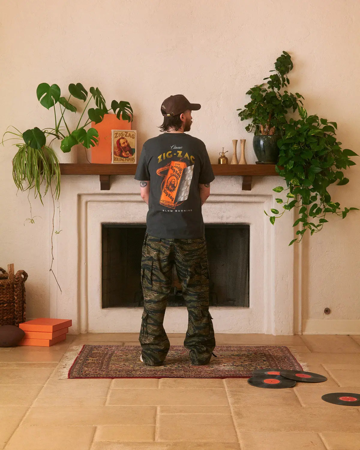 Man facing away, wearing a black Zig-Zag T-shirt with a bold orange graphic & camouflage pants, in a plant-decorated room