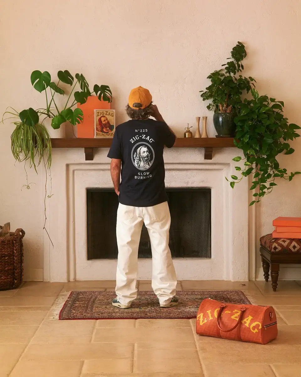 Man facing away, wearing a black Zig-Zag T-shirt and orange cap, in front of a fireplace with a Zig-Zag duffle bag nearby