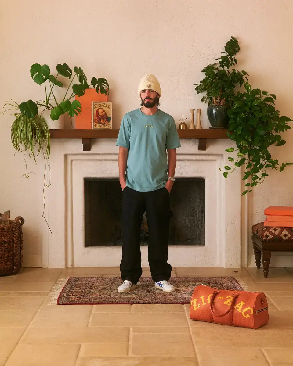 Man wearing a light blue Zig-Zag T-shirt and beanie, standing by a fireplace with a Zig-Zag duffle bag nearby