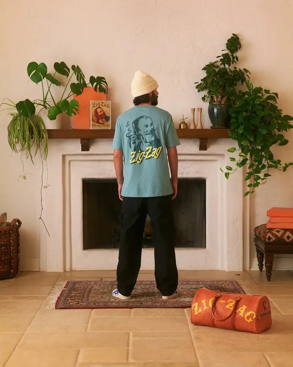 Man facing a fireplace wearing a light blue Zig-Zag shirt and beanie, with an orange Zig-Zag duffel bag nearby