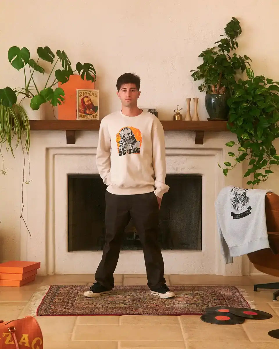 Man standing in a living room wearing a cream Zig-Zag sweatshirt featuring a graphic, surrounded by vintage decor and plants