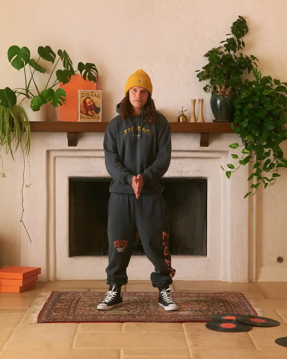 Man wearing a black hoodie, Sweatpants and yellow beanie stands in a living room with plants & a fireplace