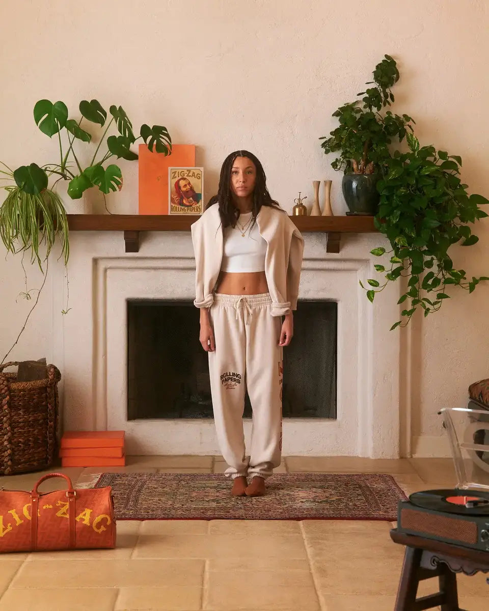 Woman wearing a casual beige outfit stands in a cozy living room with plants and a fireplace.