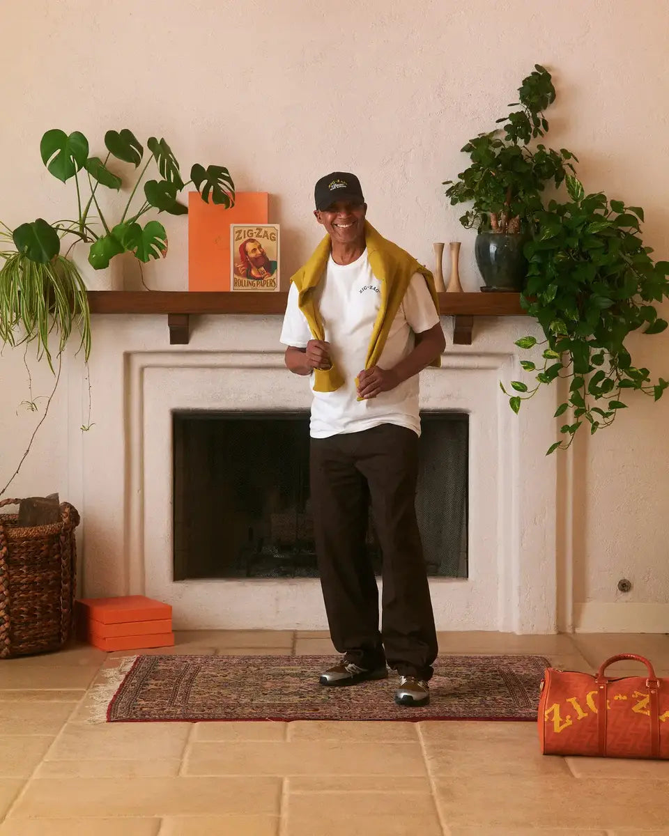 Man posing with a smile, wearing a white T-shirt, yellow sweater draped over shoulders, and a black cap, in a stylish living room