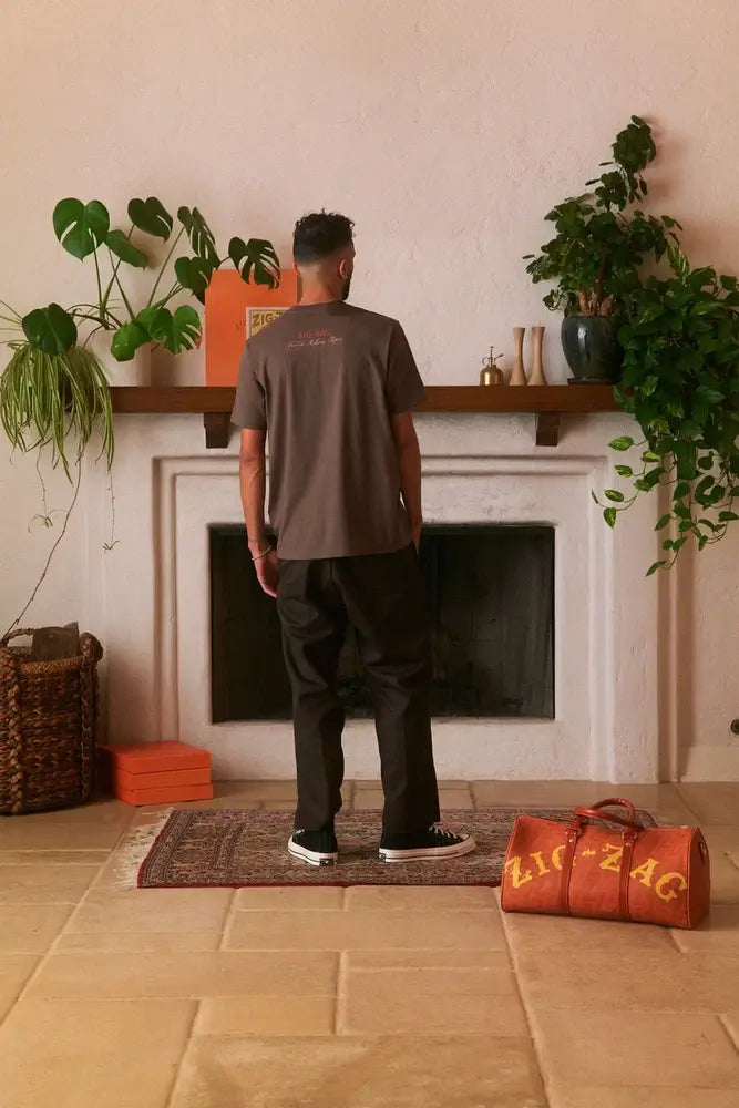 Man facing a wall, wearing a brown Zig-Zag t-shirt, standing in front of a decorated mantle and a Zig-Zag duffel bag