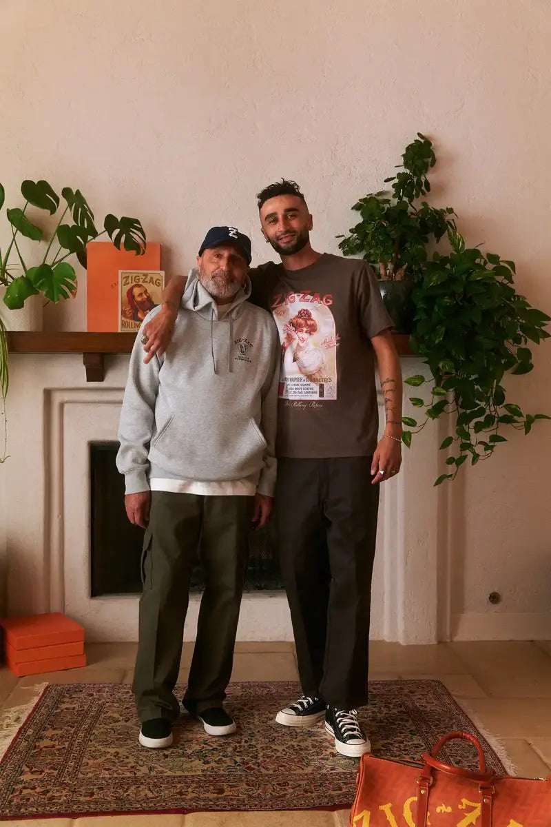 Two men standing in a living room, one wearing a gray hoodie & the other a brown Zig-Zag graphic t-shirt, both smiling