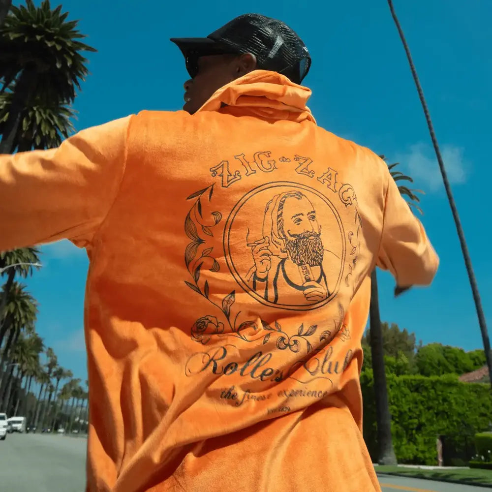 Man wearing a black cap and an orange Zig-Zag hoodie with an illustrated design and branding on the back, outdoors under a blue sky