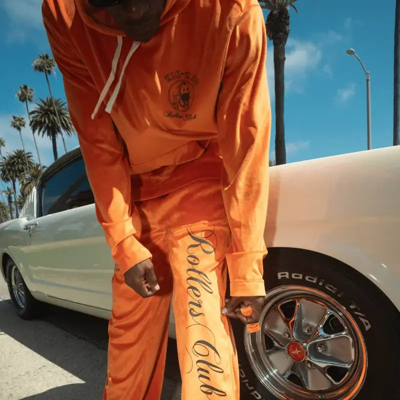 Man dressed in an orange "Zig-Zag Rollers Club" outfit leaning casually against a classic white car, with palm trees in the background