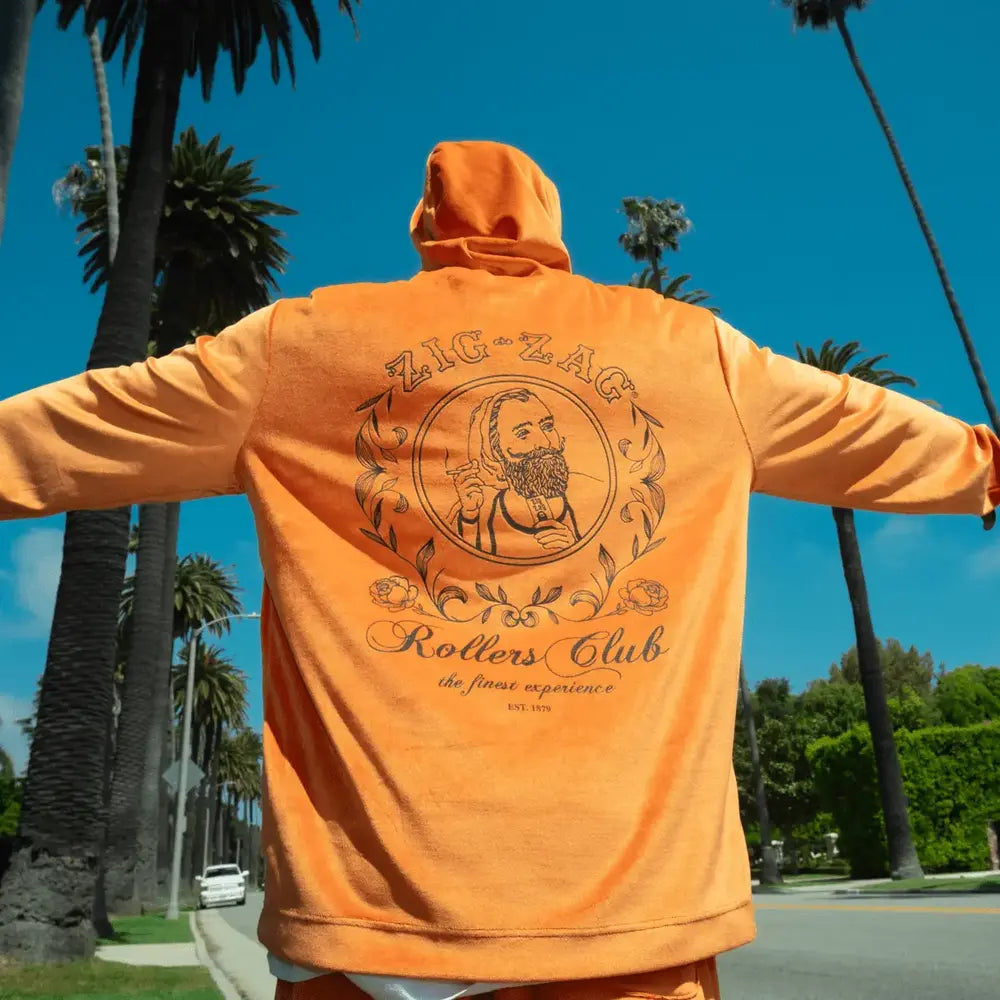 Man wearing an orange Zig-Zag "Rollers Club" hoodie with arms outstretched, standing under palm trees against a blue sky