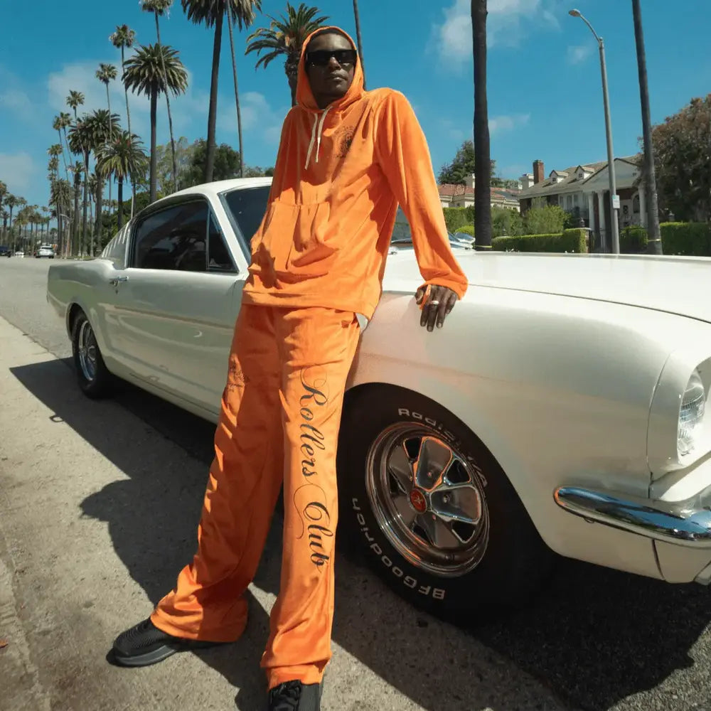 Man in an orange "Rollers Club" hoodie and pants leaning against a classic white car, under palm trees